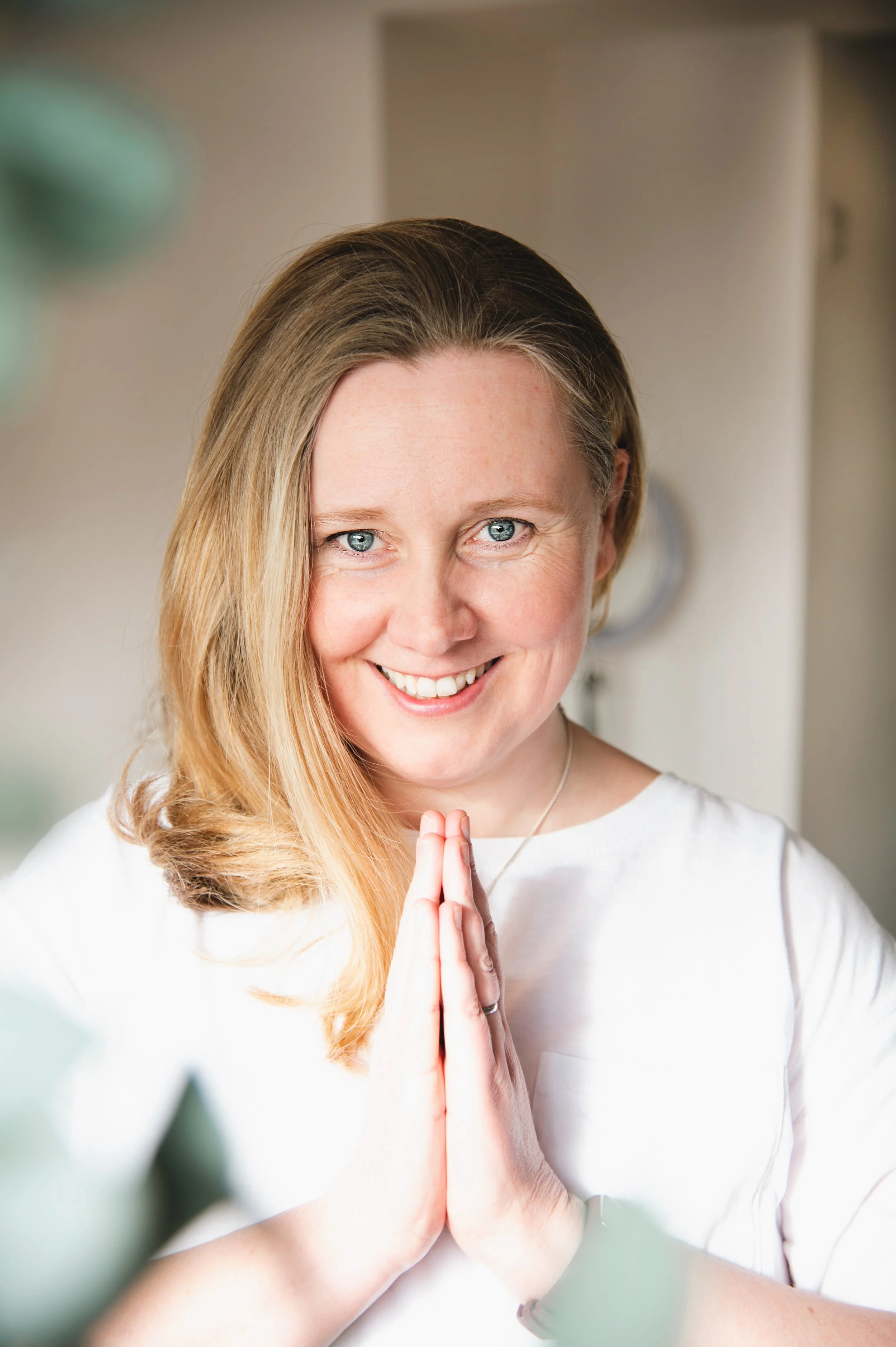 A woman with blonde hair and blue eyes smiling softly with her hands pressed together in a prayer or meditation pose, wearing a white top, standing indoors with a blurred background.