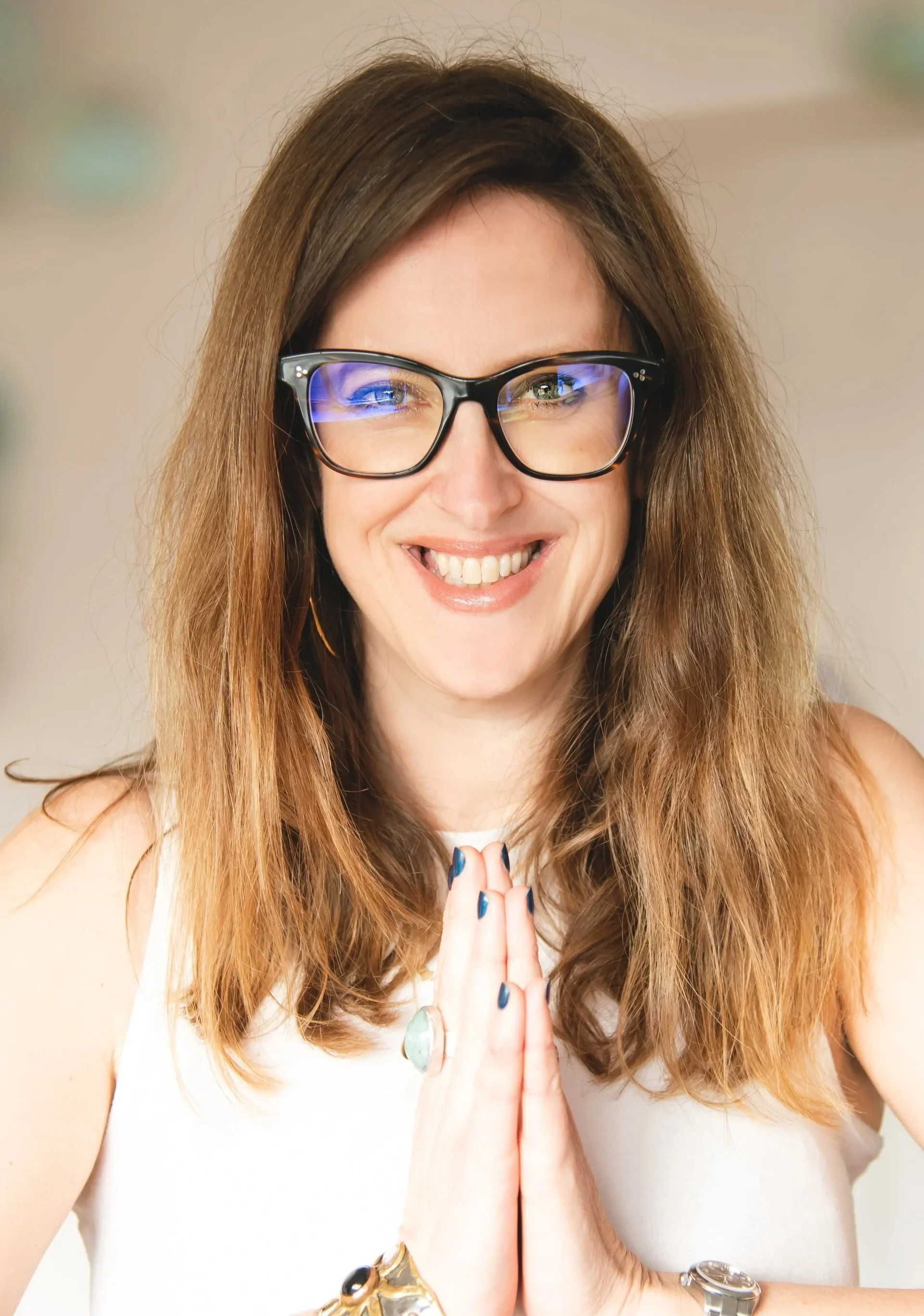 A Reiki practitioner with long brown hair, wearing glasses, smiling, holding her hands together in a praying gesture, wearing a white top, and jewelry on her wrists. Shot by Wellness Brand Photographer, Emily Dyer Creative