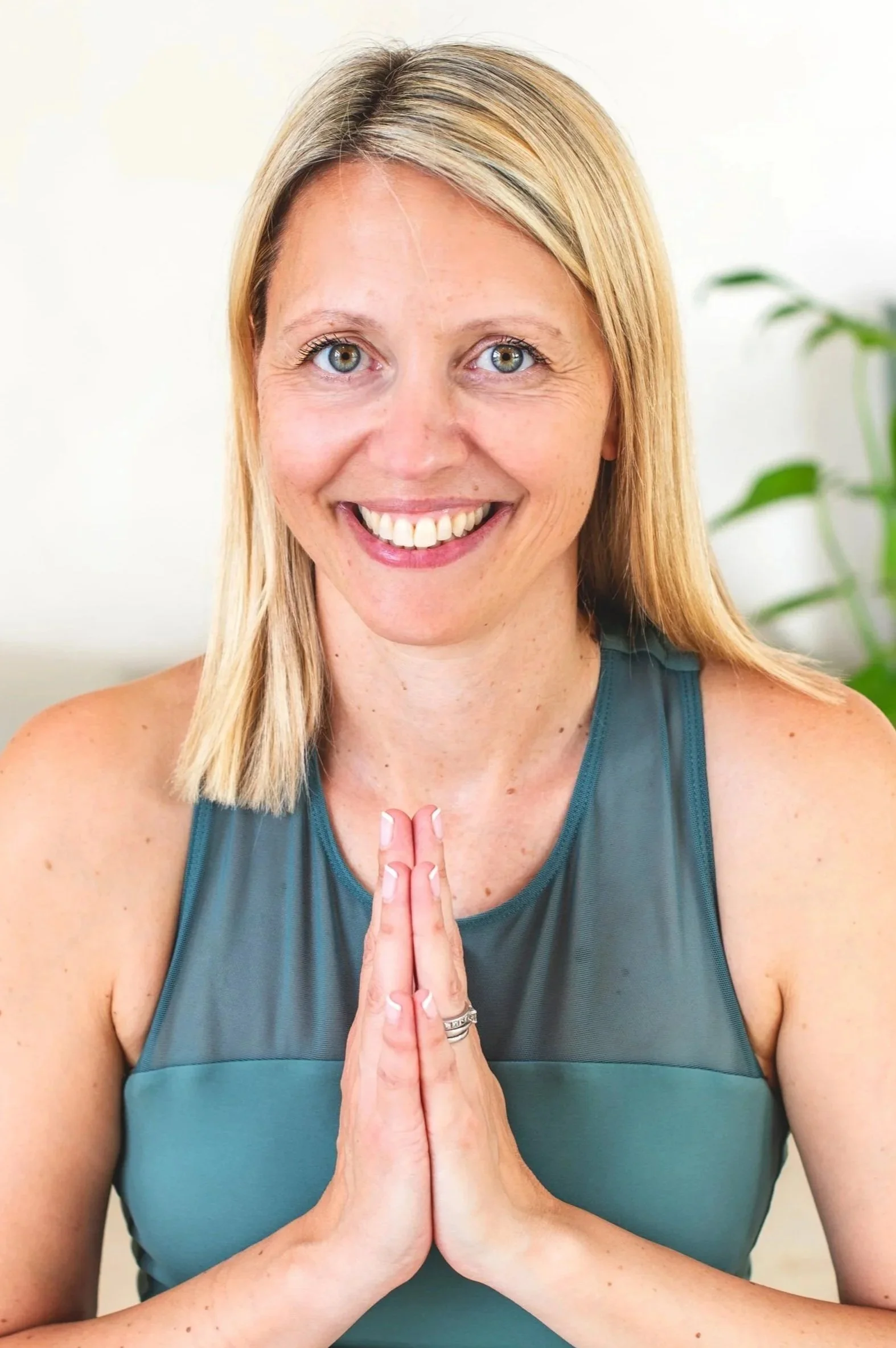 A woman smiling with her hands pressed together in a prayer pose, wearing a teal athletic tank top, with a background of a white wall and a green plant.