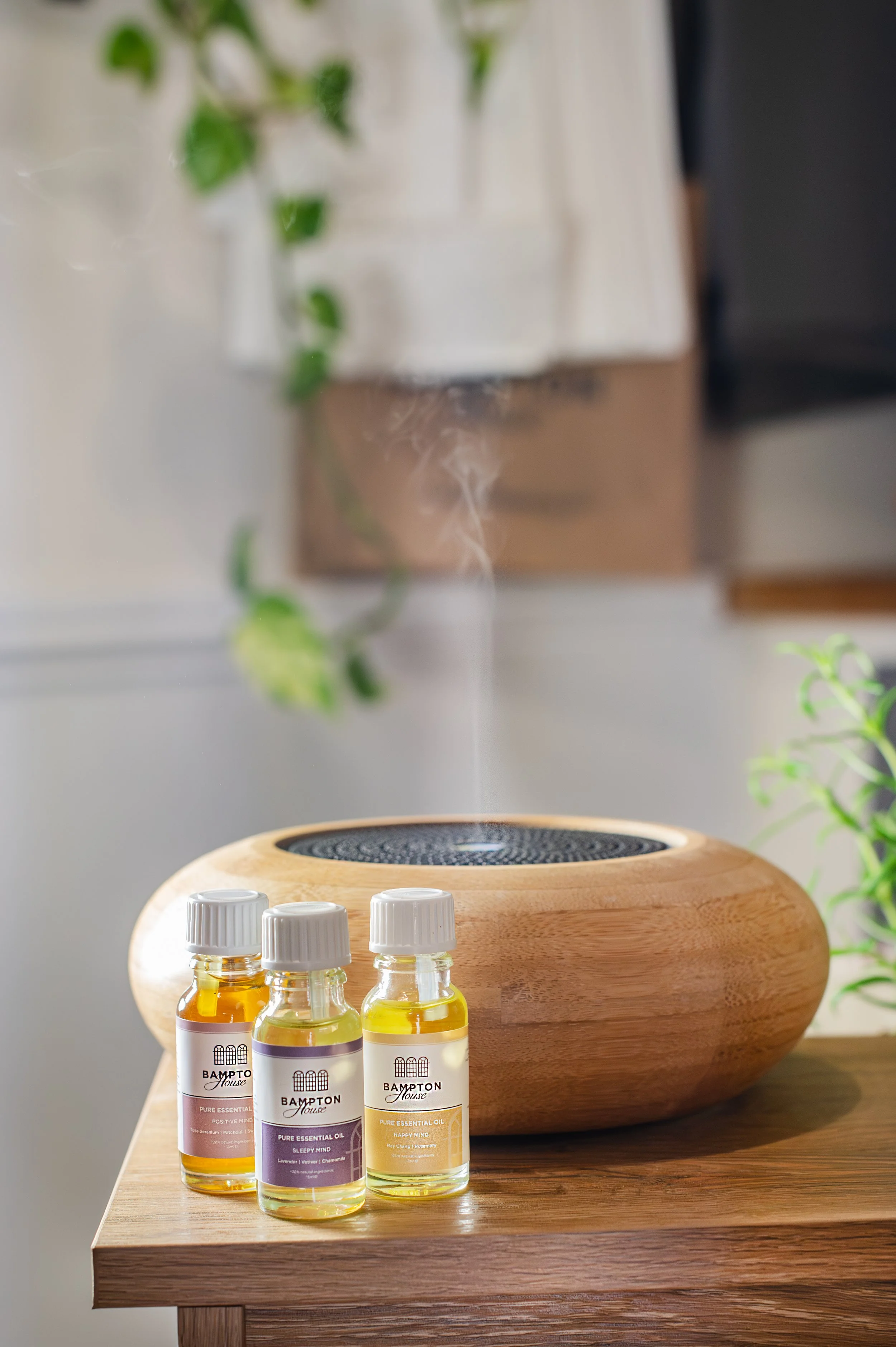 Three bottles of Bampton House essential oil on a wooden table, a large wooden diffuser behind them, and a white vase with greenery in the background. Shot in Witney, Oxfordshire, by Wellness Brand Photographer, Emily Dyer Creative