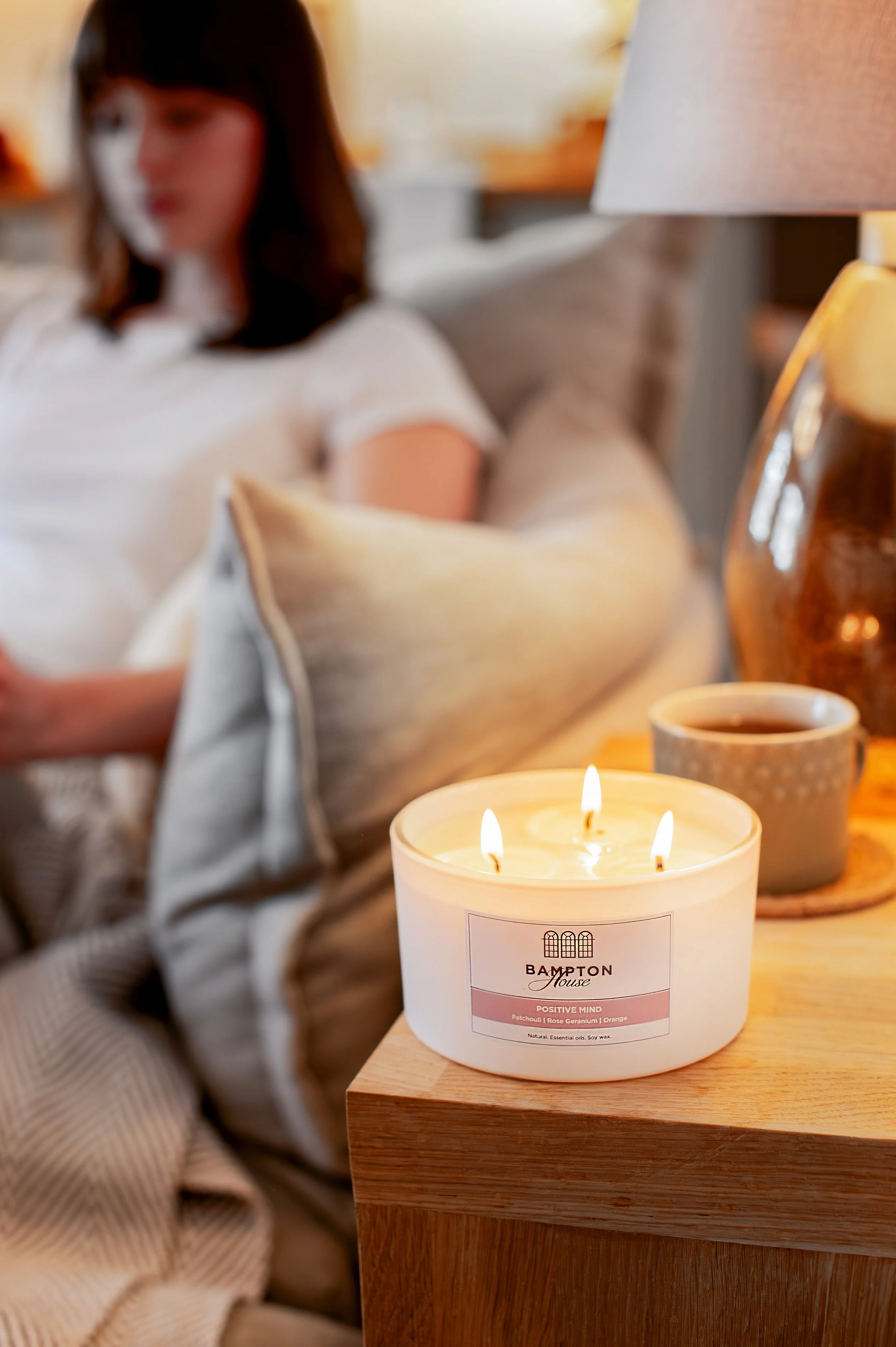 A lit white candle on a wooden table labeled 'Bampton House,' with a woman sitting in the background, blurred, in a cozy indoor setting. Shot in Witney, Oxfordshire, by Wellness Brand Photographer, Emily Dyer Creative