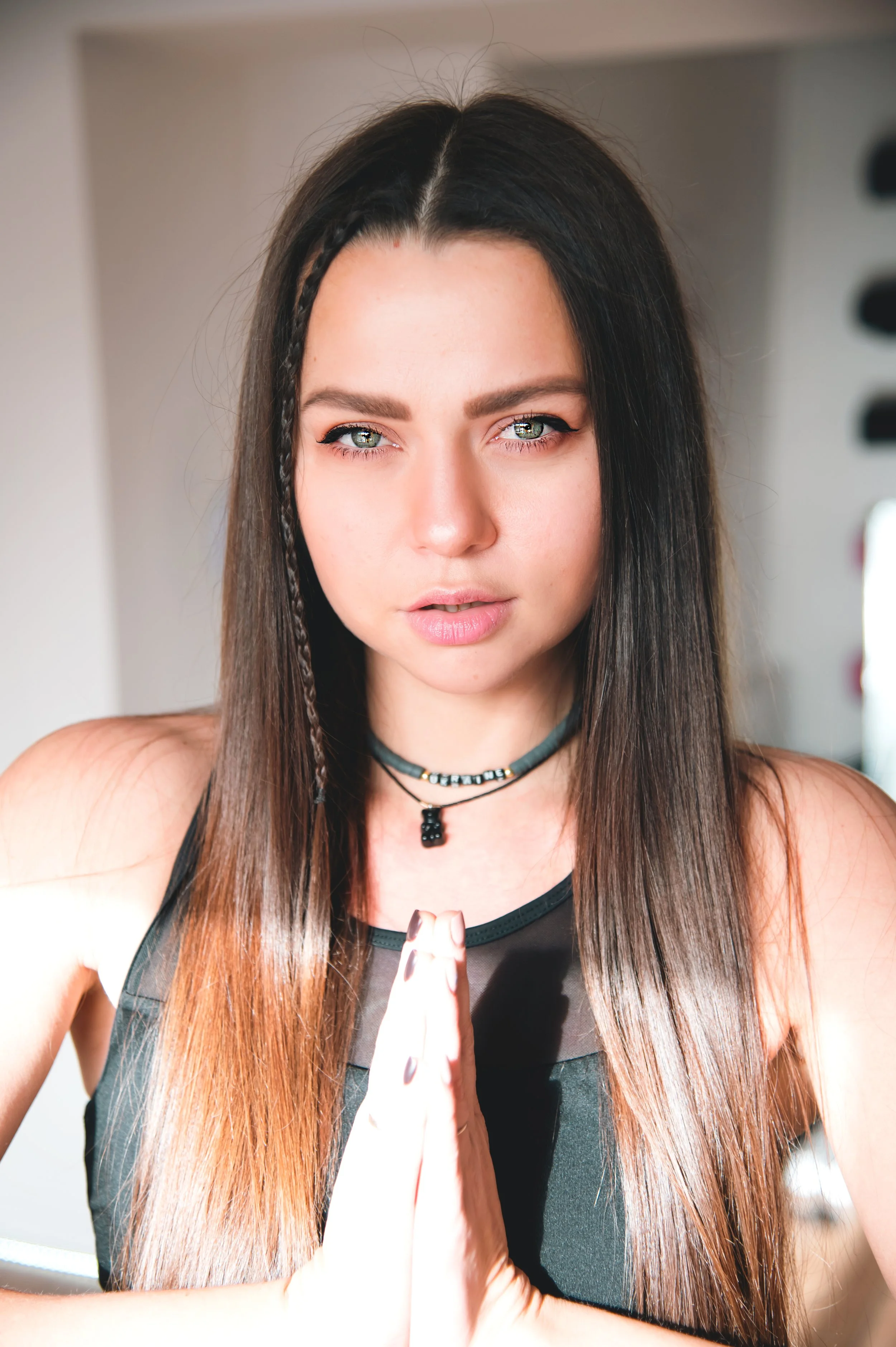 A young woman with long brown hair and blue eyes, wearing a black sleeveless top and layered necklaces, is seen in a close-up photo with her palms pressed together in front of her chest, looking directly at the camera.