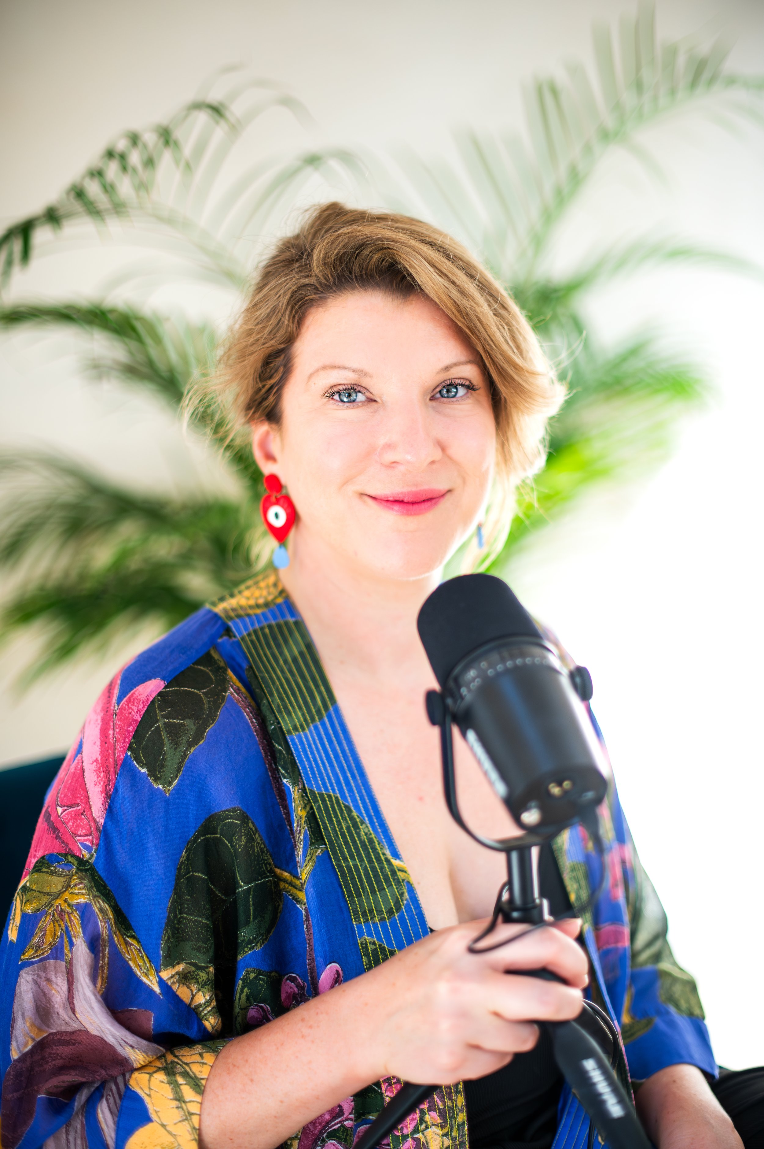 A woman with short wavy hair, blue eyes, and colorful earrings smiling and holding a microphone. She is wearing a multicolored, patterned top and sitting in front of a large leafy plant.