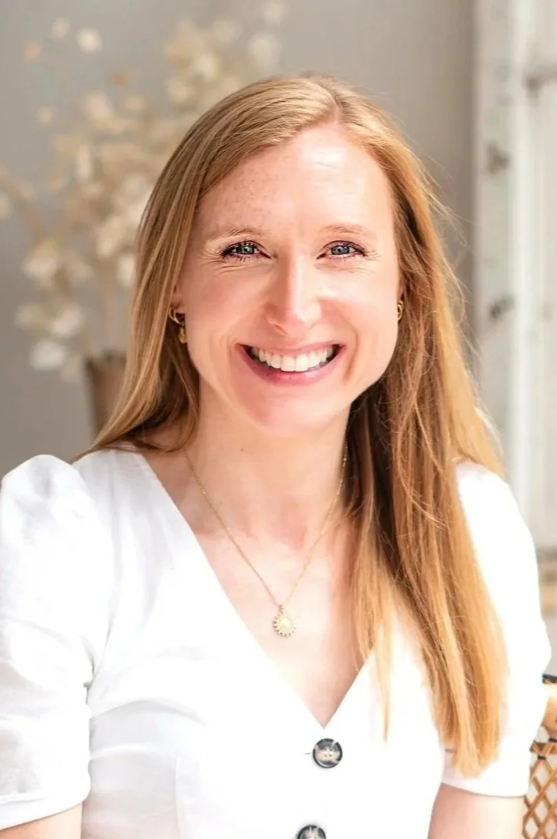 A yoga teacher, smiling woman with red hair wearing a white top and gold jewelry, sitting in a bright room with a blurred background. Shot by Wellness Brand Photographer, Emily Dyer Creative