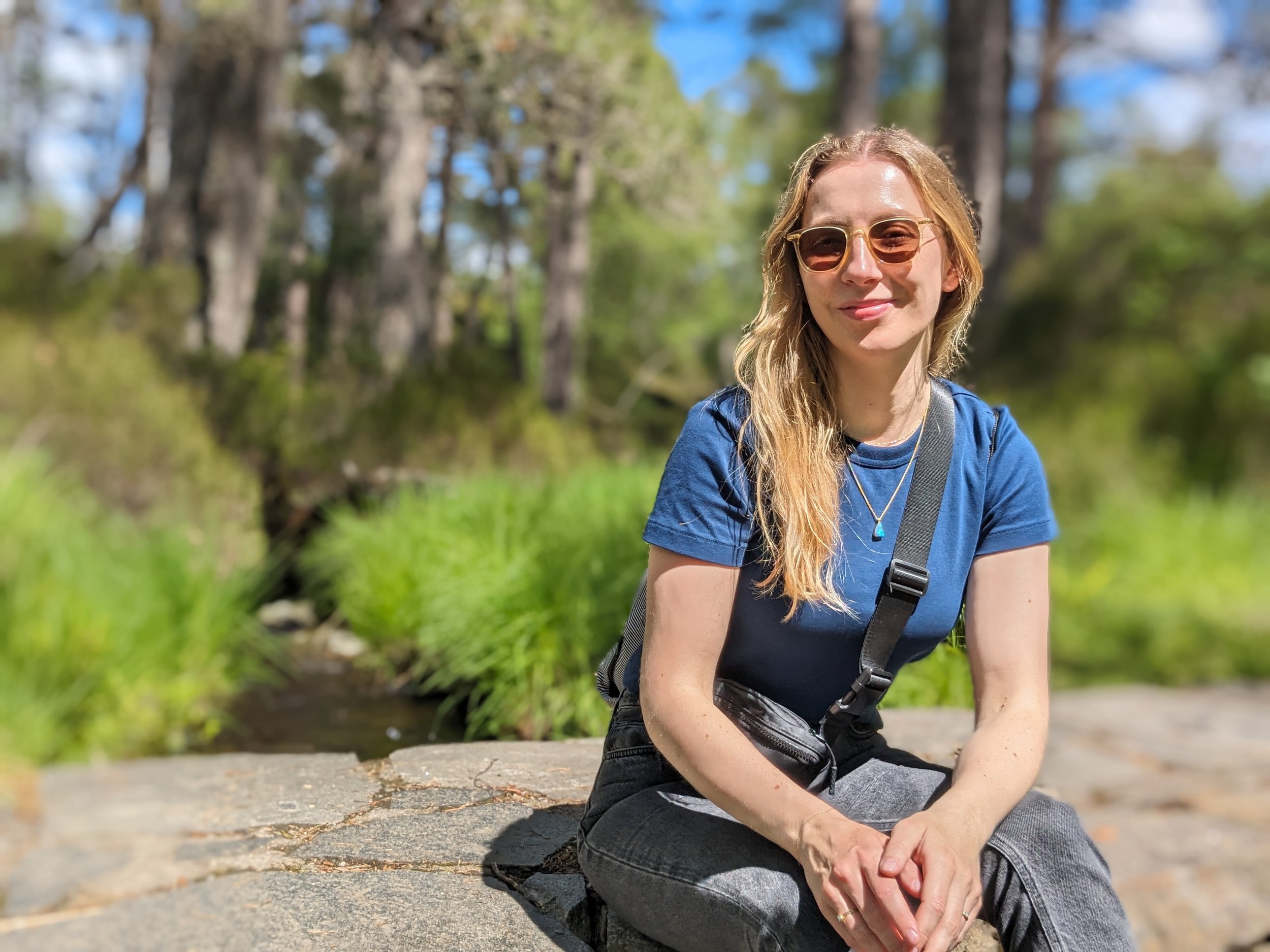 Stefanie Maier mit Sonnenbrille und blauer Kleidung sitzt draußen auf einem Felsen, umgeben von grüner Natur und Bäumen, bei sonnigem Wetter.