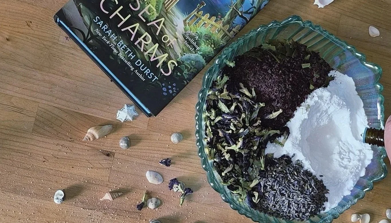 Bowl of bath salt that is not mixed yet with the Sea Of Charms book by Sarah Beth Durst in the background among sea shells