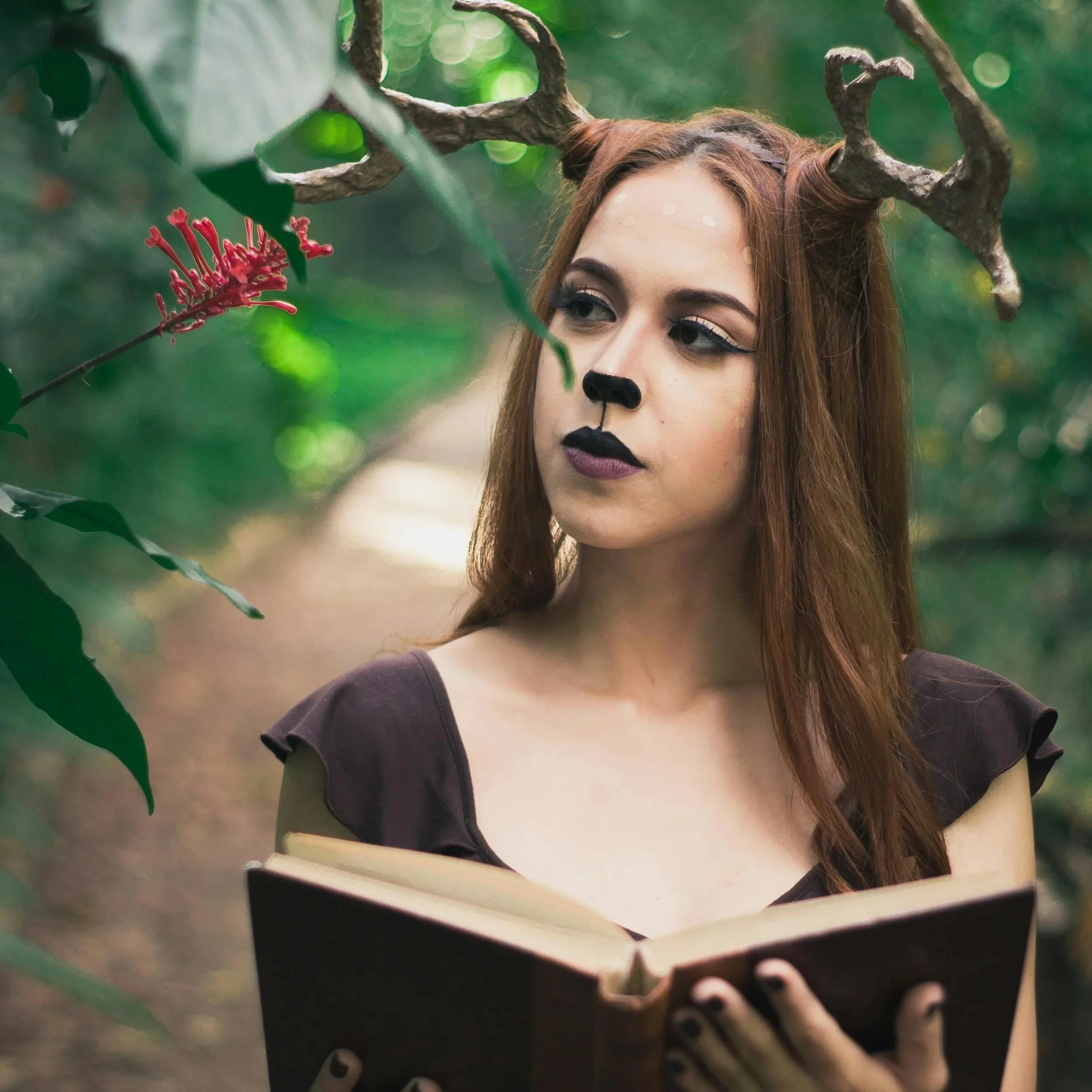 A woman with deer antlers and black makeup, including nose and lips, stands in a forest holding a book.