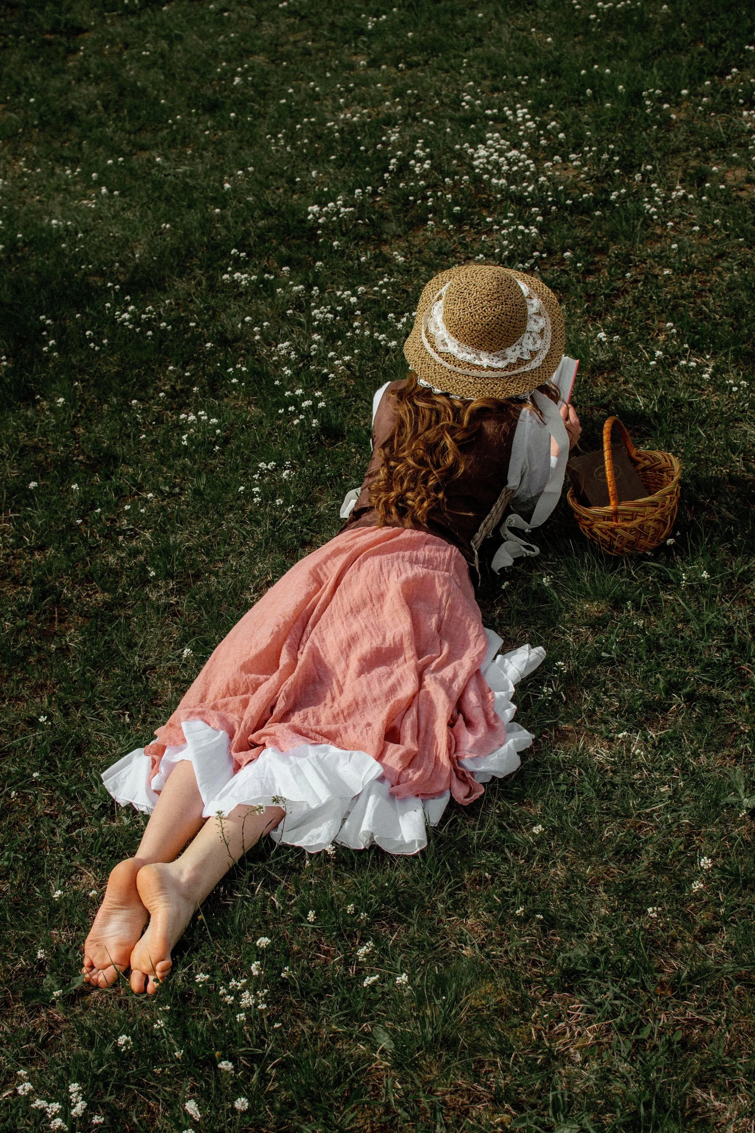 Person in a sunhat and skirt laying on their stomach and reading outside with a basket