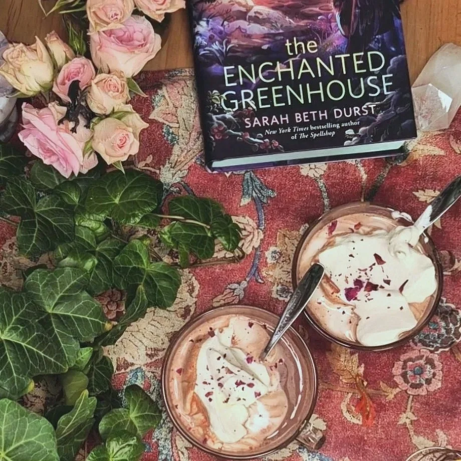 A table with a floral tablecloth, featuring two glasses of rose hot chocolate with whipped cream, a bunch of pink and white roses, a potted ivy plant, and a copy of the book 'The Enchanted Greenhouse' by Sarah Beth Durst.