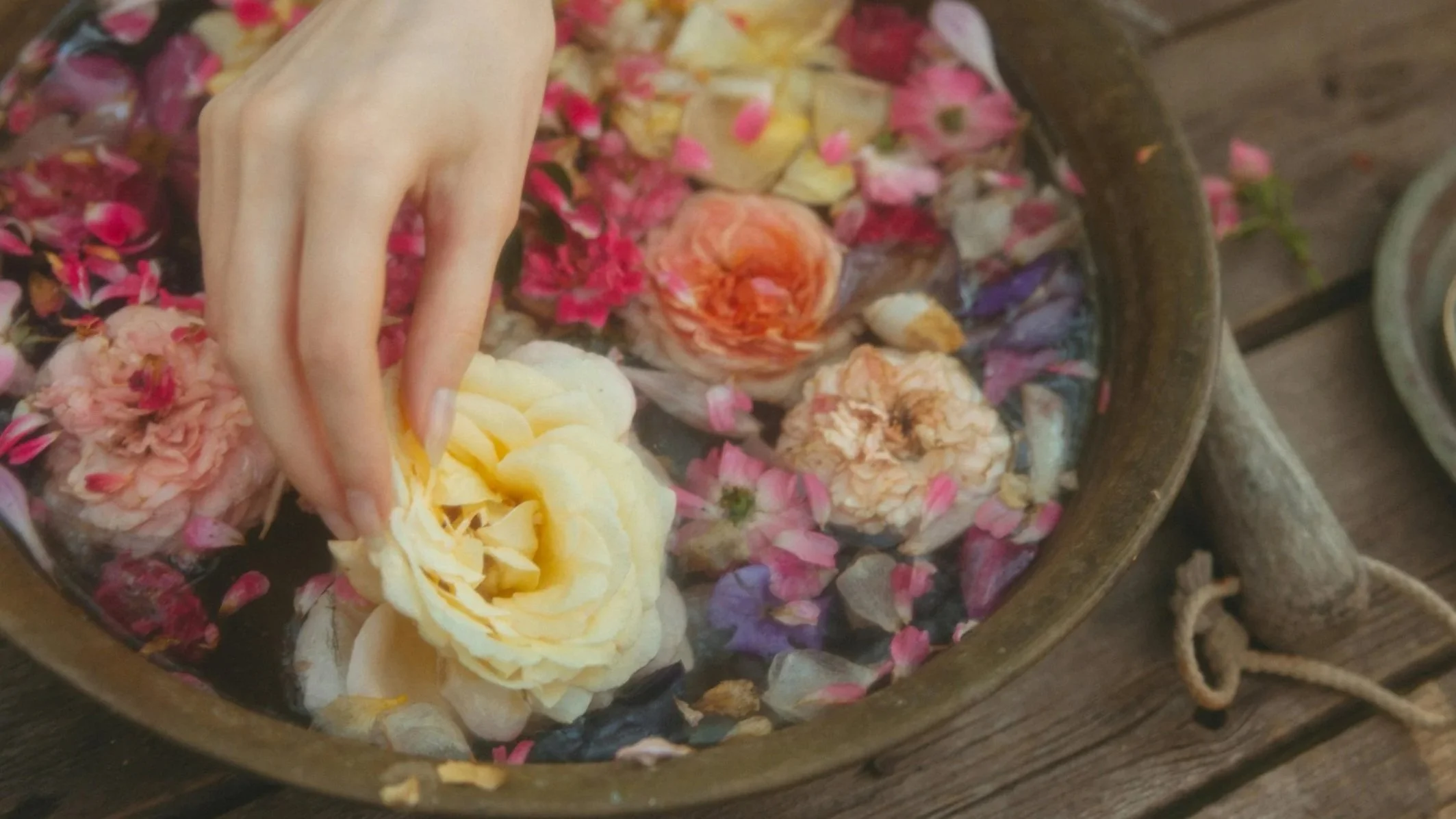 flowers floating in a bowl of water on a wooden table with a hand reaching in to touch one of the flowers