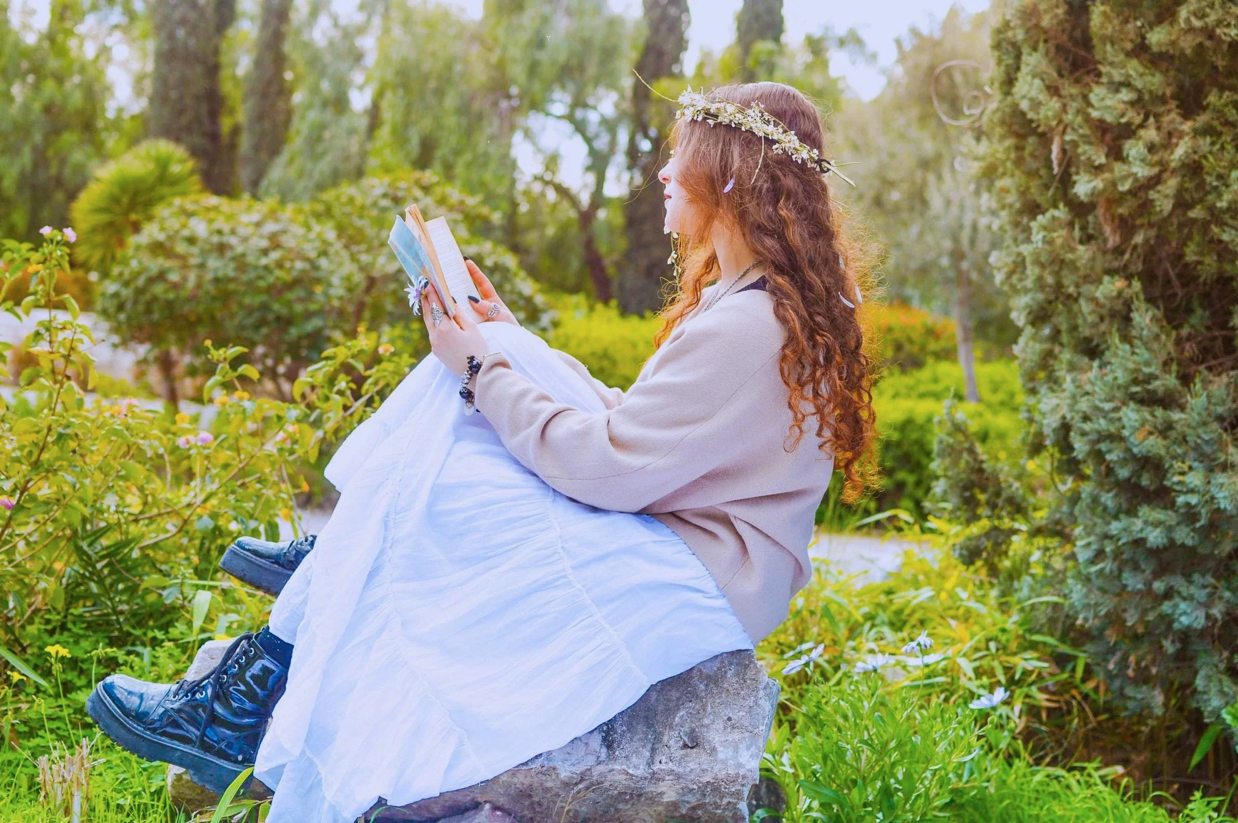 A young woman with long curly hair, wearing a flower crown, sitting on a rock in a lush green garden, reading a book.