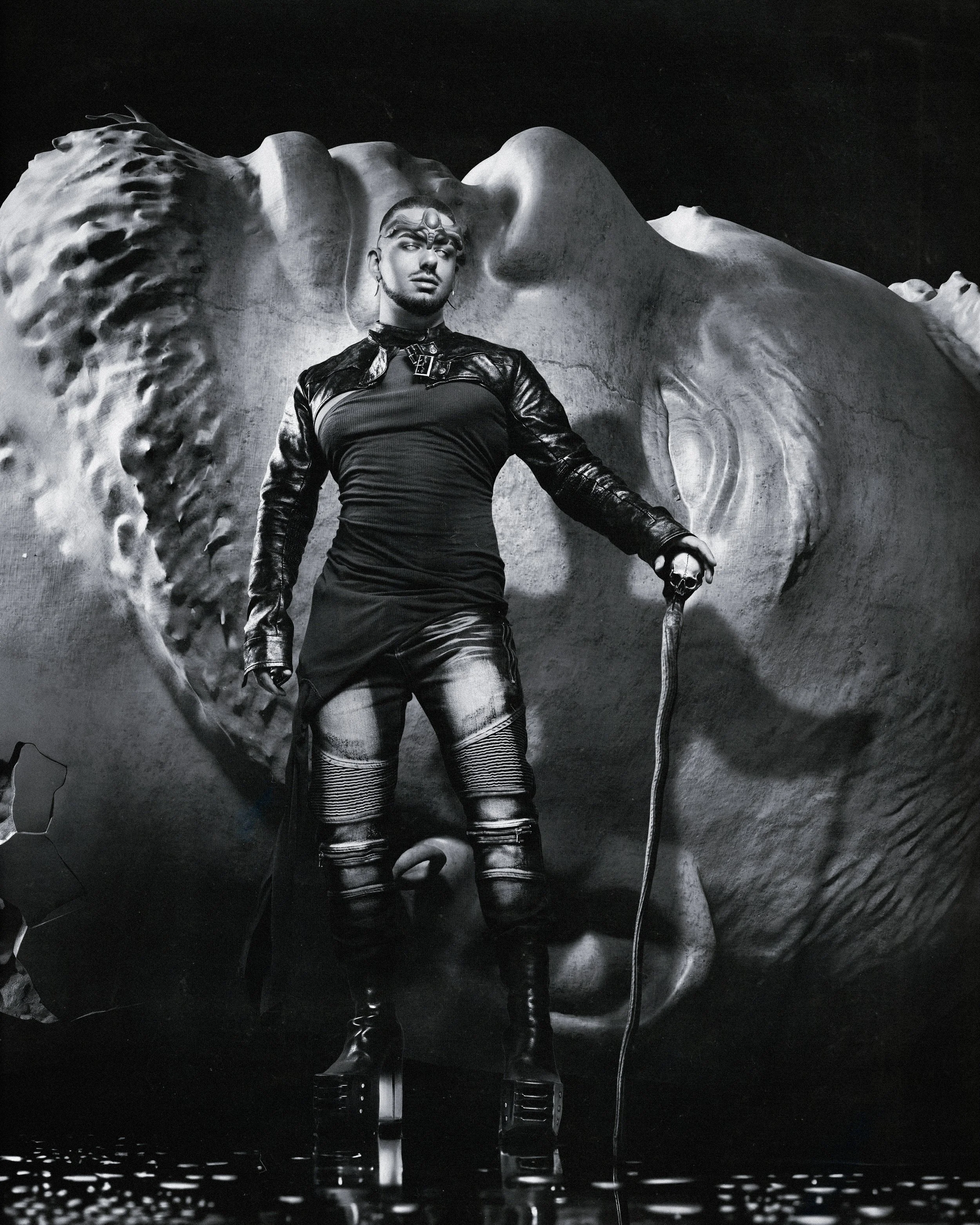 A person, David Oldenburg,  dressed in a black leather outfit with high boots posing in front of a large face sculpture. The person holds a staff with a skull on top, and the scene is in black and white.