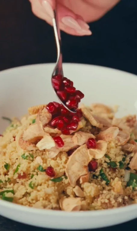 A bowl of couscous topped with chicken, mushrooms, and pomegranate seeds, with a hand holding a spoon above it.