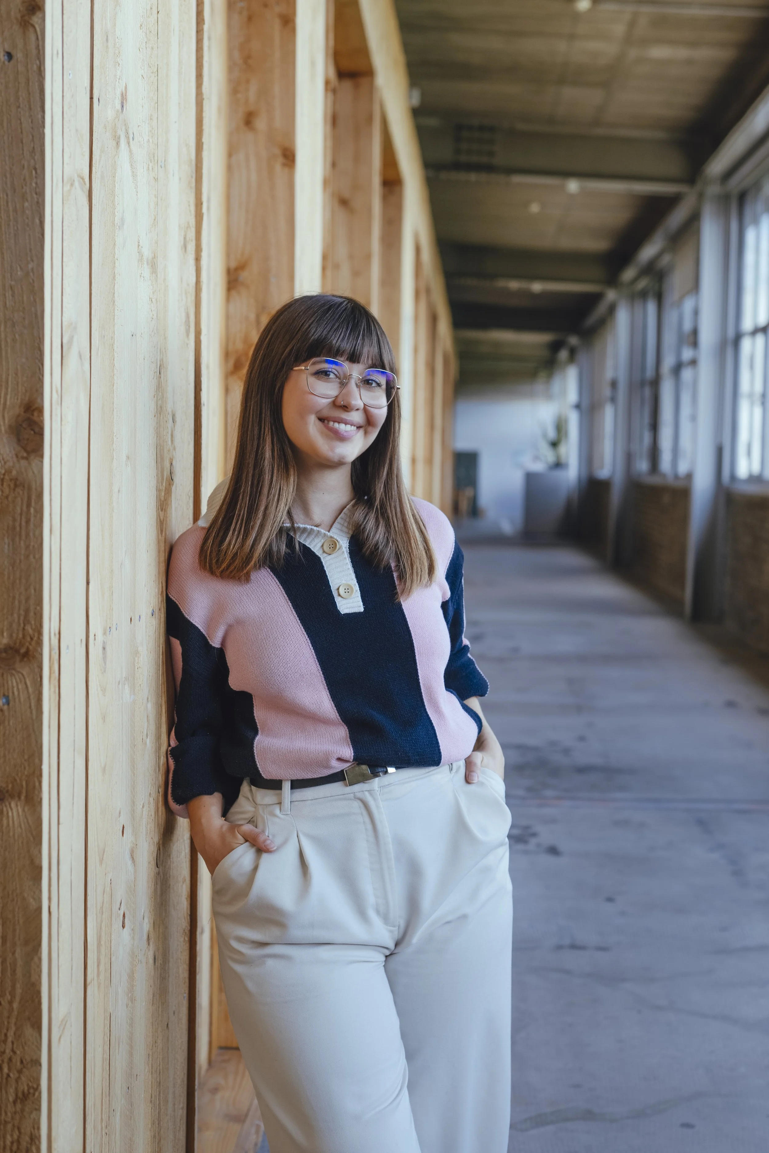 A young woman with glasses and shoulder-length brown hair, smiling and leaning against a wooden wall inside a building with large windows and a wooden ceiling.