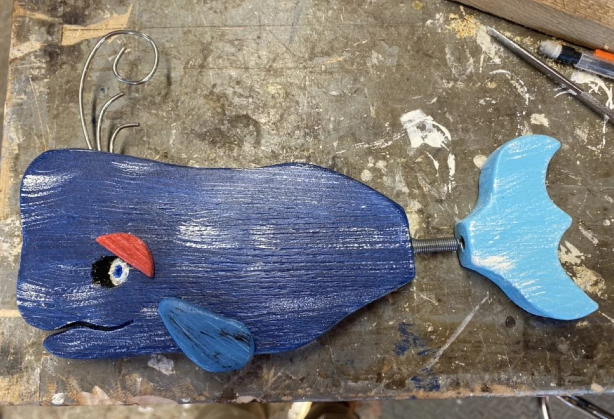 Handcrafted wooden whale with painted features, resting on a workbench with tools and materials around it.