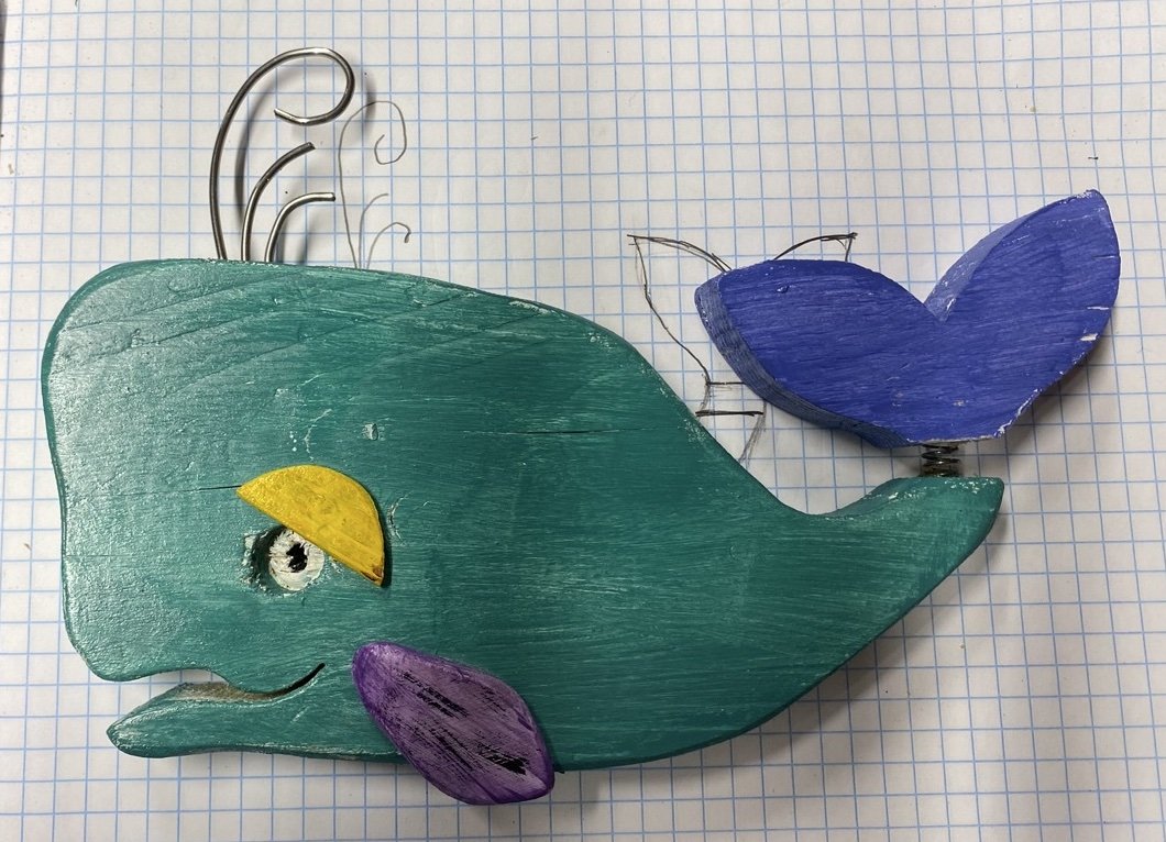 Colorful wooden whale craft with purple and blue fins, yellow eye, and decorative wire spout on graph paper background.