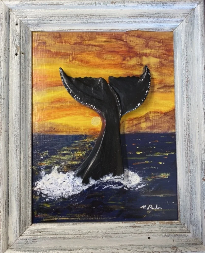 A wall-mounted sculpture of a whale tail emerging from the ocean, set in front of a sunset over the water, framed with white painted wood.