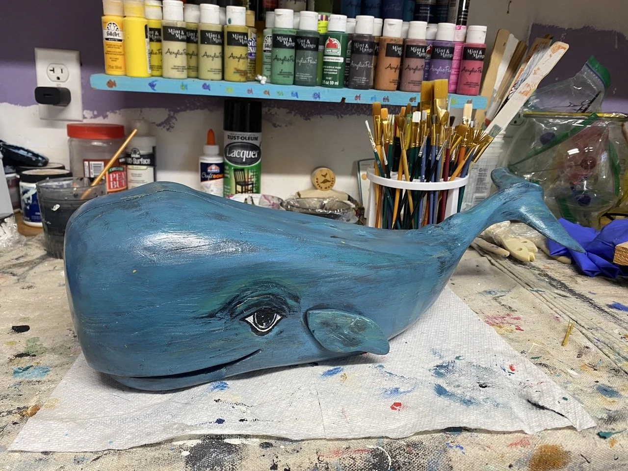 A painted whale sculpture with a blue body, decorated with a detailed eye and a gentle smile, on a cluttered artist's work table.