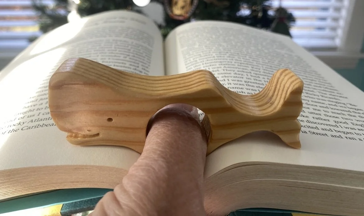 A wooden whale resting on an open book with a person's finger through the  loop, in front of a Christmas tree.