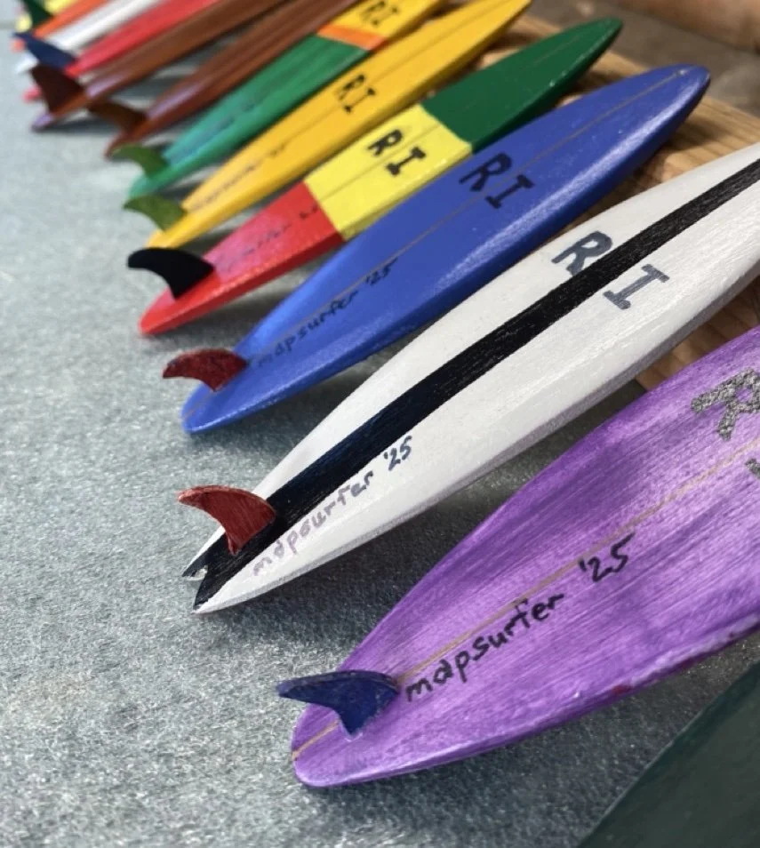 Colorful wooden surfboard-shaped objects, each painted in different colors including purple, white, blue, green, yellow, red, and brown, labeled with 'map surfer ' and a year, arranged diagonally on a gray surface.