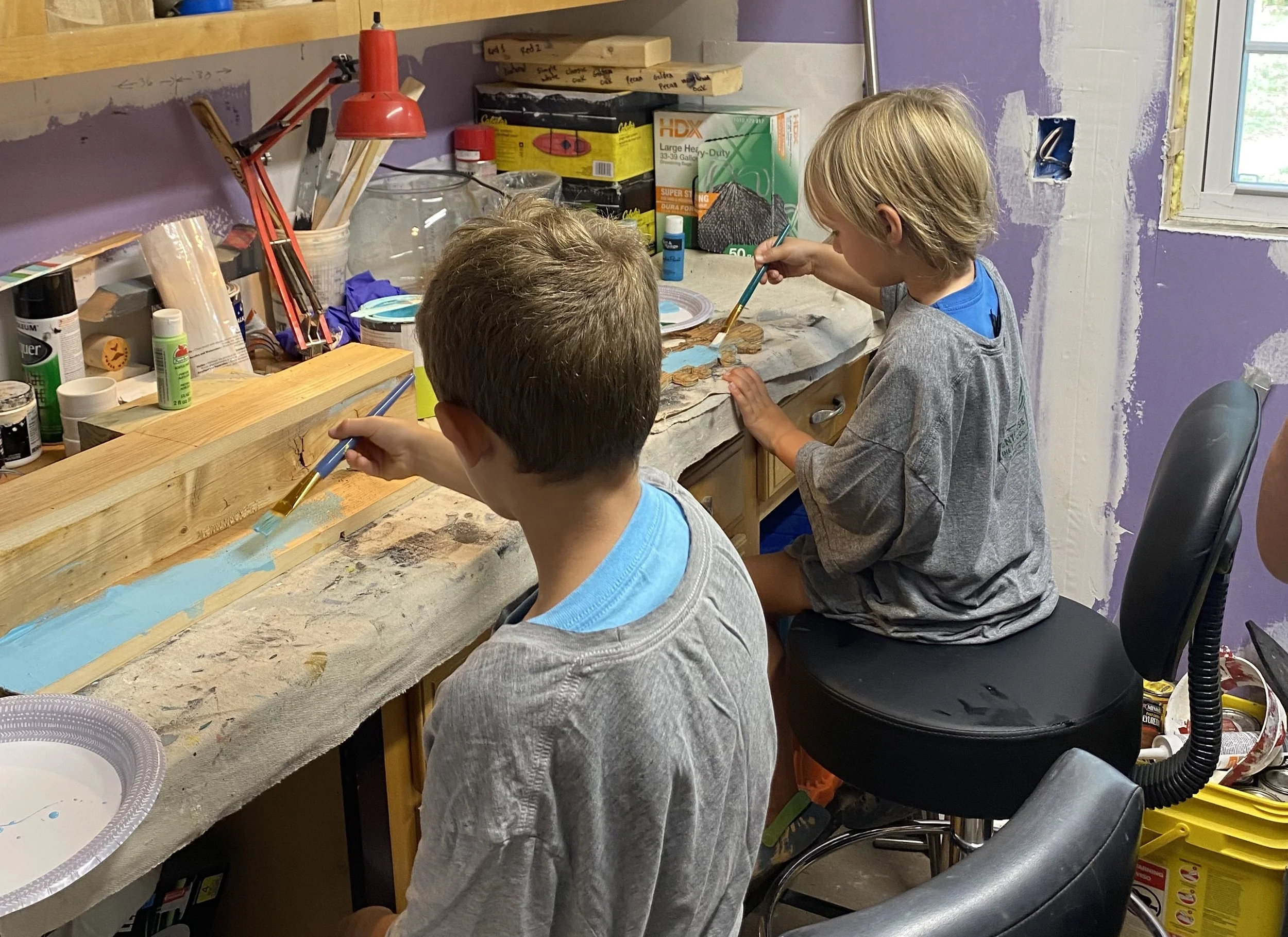 Two children painting on a long piece of wood in a workshop or craft room.