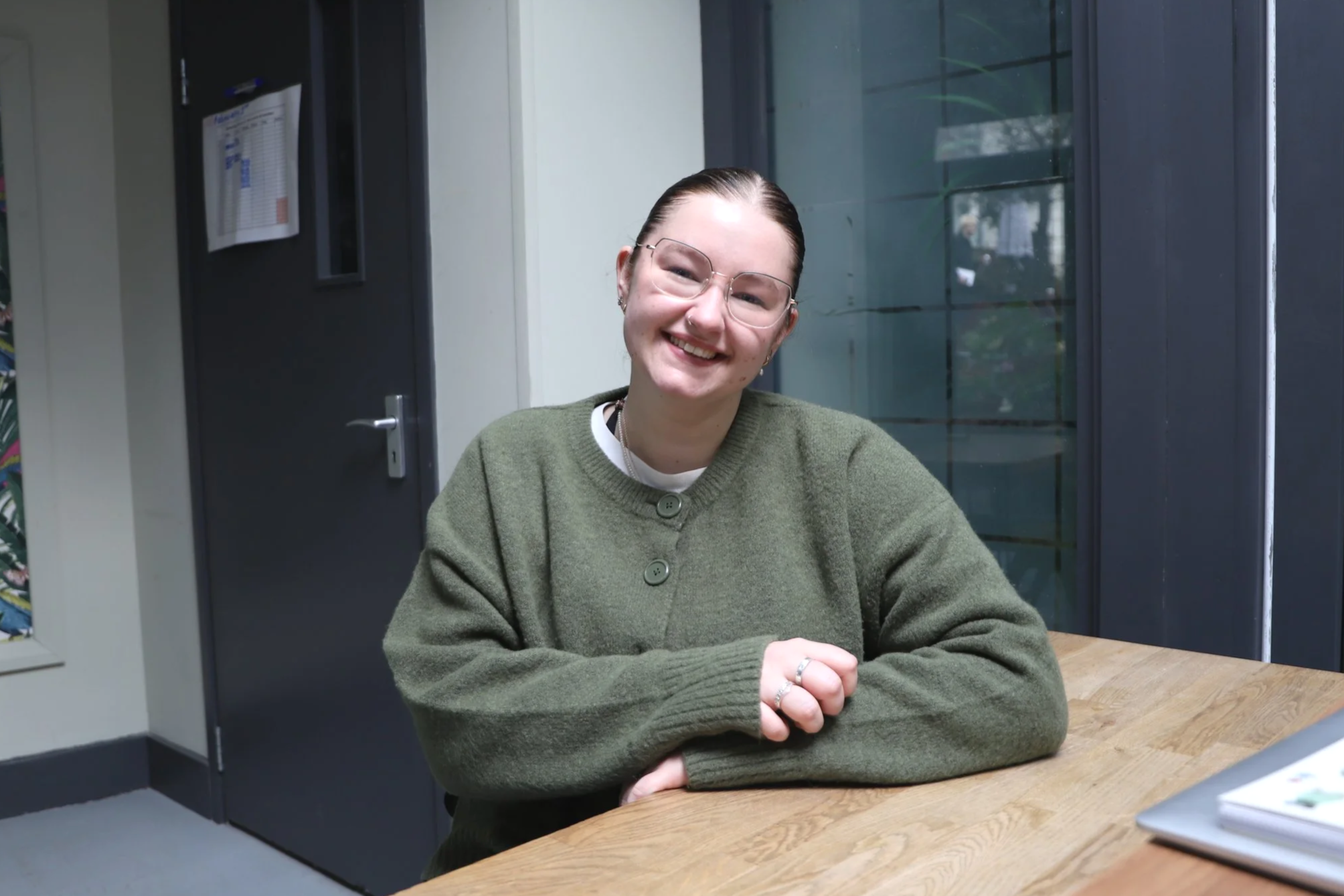 A woman with slick back brown hair smiles while sitting at a wooden desk with a silver laptop, a window and a door to a separate room in the background