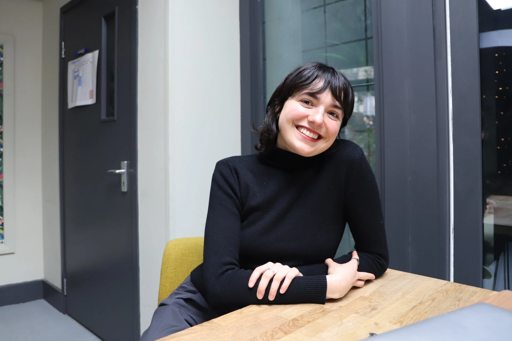 Woman with short dark hair wearing a black long sleeve jumper, sitting at a wooden desk. In the background, there is a framed tropical picture, a window and a door that leads to another room