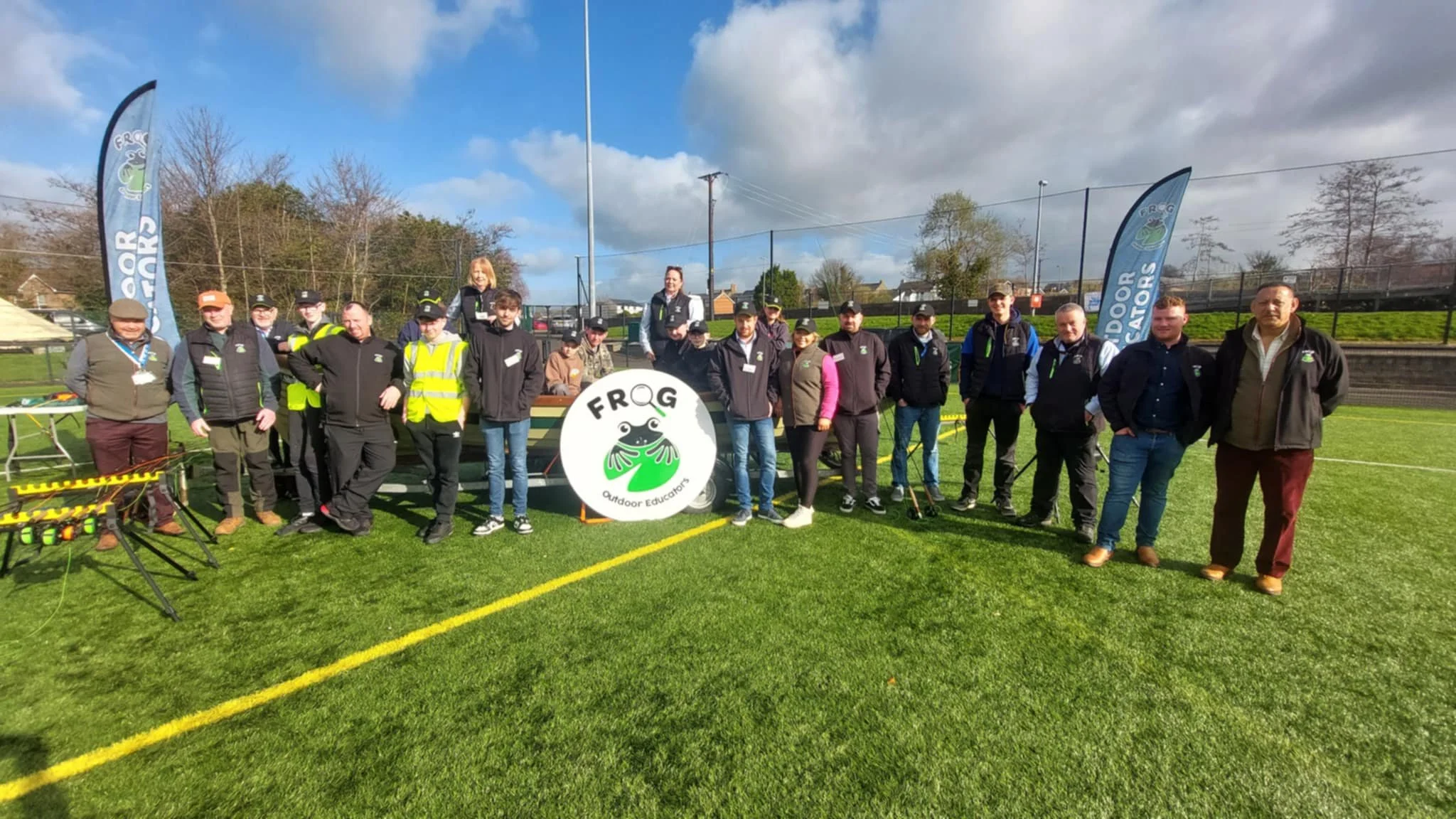 Group of people standing on a sports field with flags, banners, and a round sign that reads 'FROG Outdoor Educators' featuring a frog logo, under partly cloudy skies.