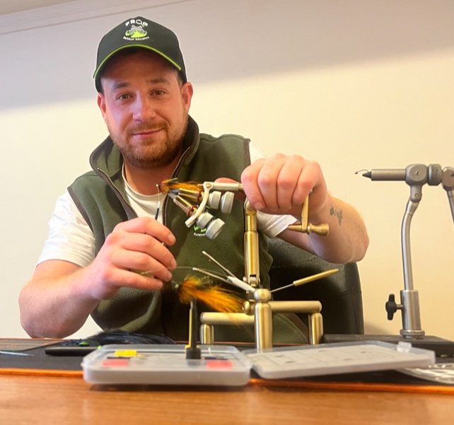 Man sitting at a desk creating a fishing lure using fly tying tools and materials.