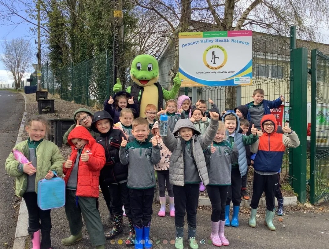 Group of children and a mascot standing in front of a Dennett Valley Health Network sign outside, dressed in rain gear and rain boots, smiling and giving thumbs up.