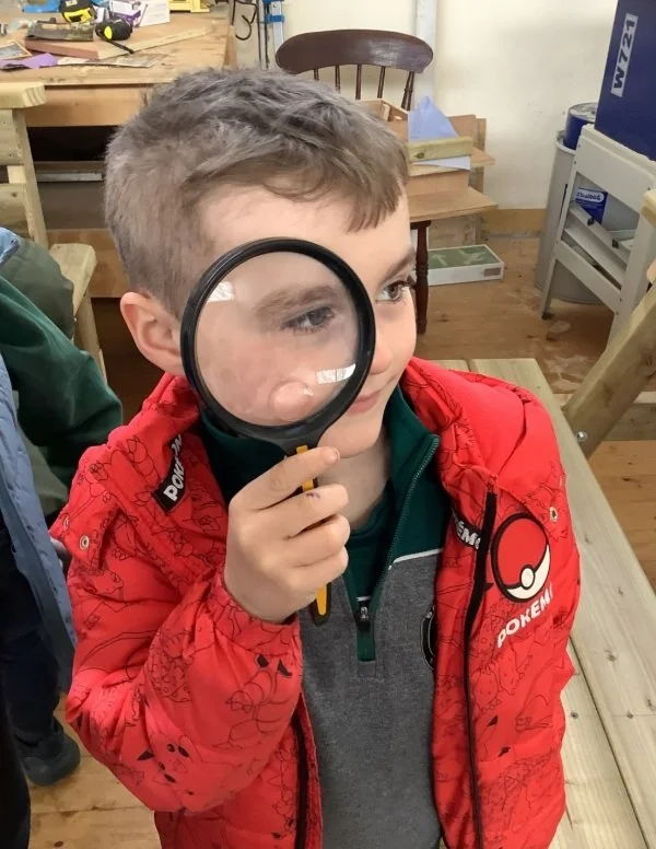 Young boy wearing a red Pokémon jacket holds a magnifying glass to his left eye, looking intently at something outside the frame in a workshop or classroom setting.