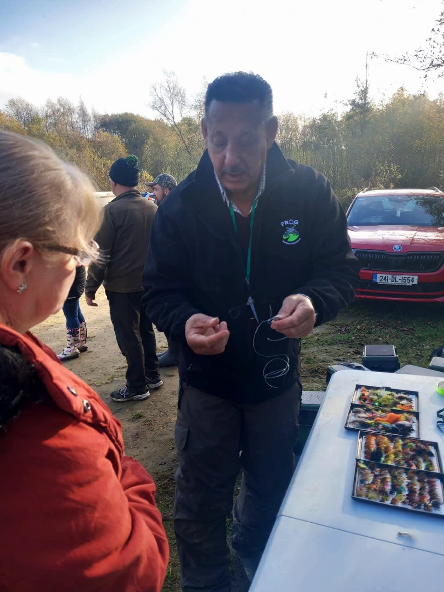 A man in a black jacket with a logo talking to a woman in a red jacket at an outdoor event with zoomed fly fishing flies on display.