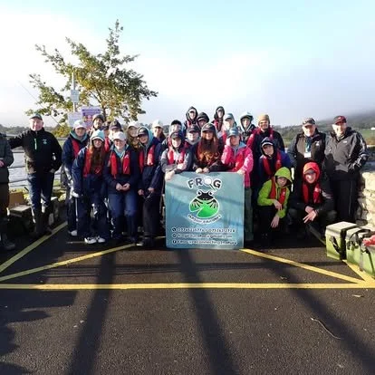 Group photo of people with backpacks and hats holding a banner with a frog logo, outdoors in a parking lot on a sunny day.