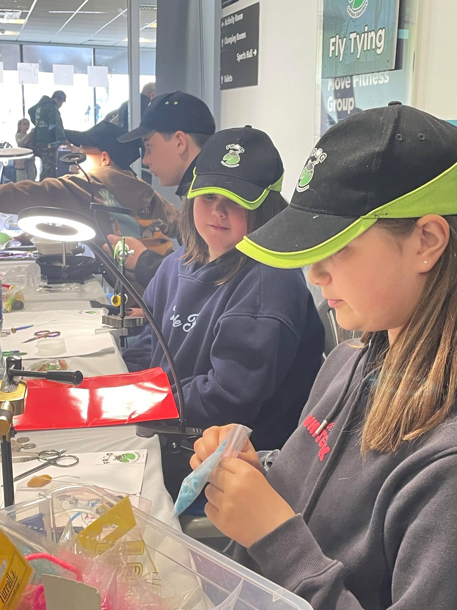 Three young girls at a crafting table, all wearing black caps with a logo, working on fly-tying at a workshop. In the background, other children and a sign for a fly-tying and fitness group can be seen.