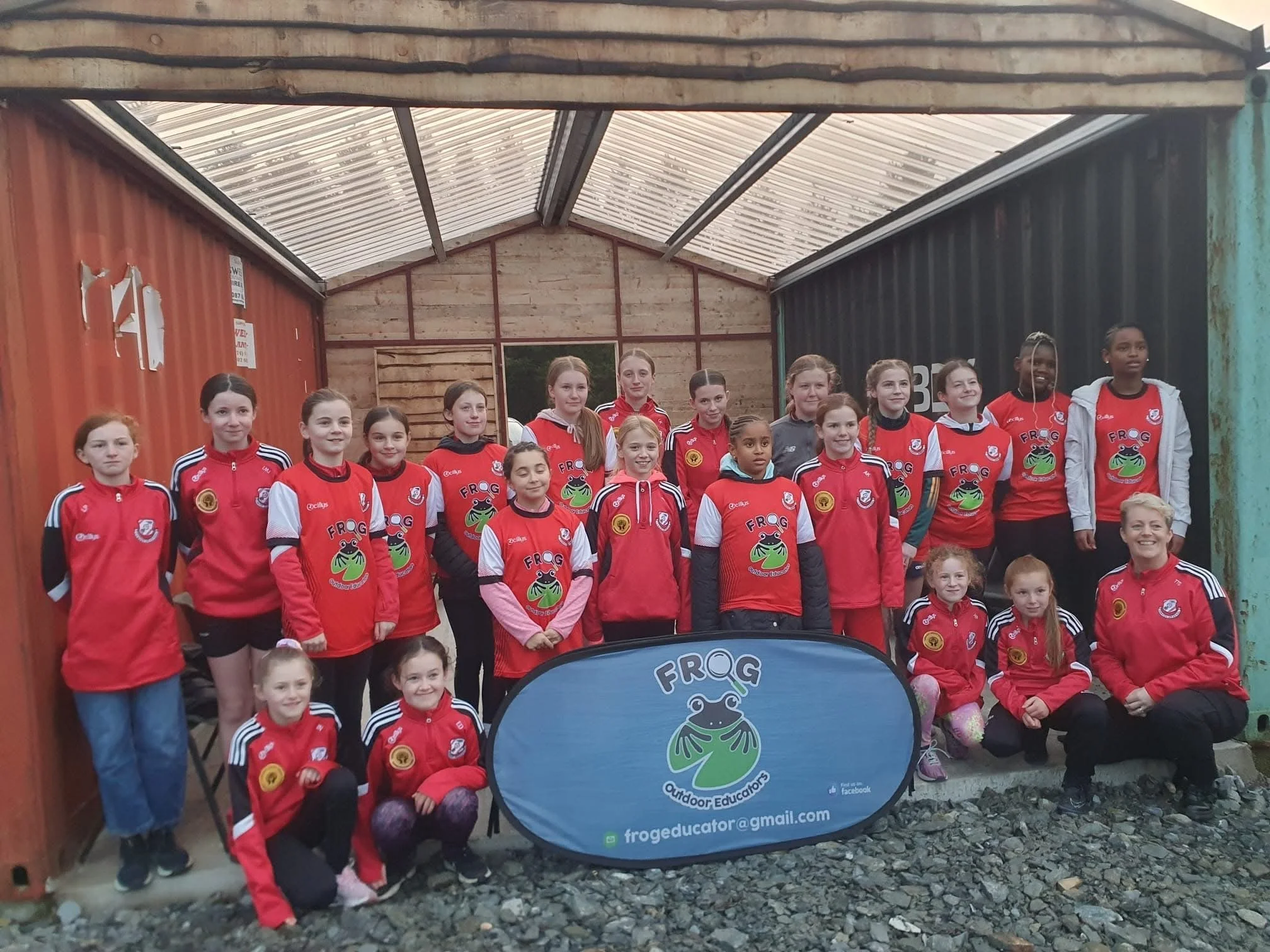 Group of young girls in red sports jerseys and jackets posing inside a wooden and metal structure with a blue Frog Educators banner in front.