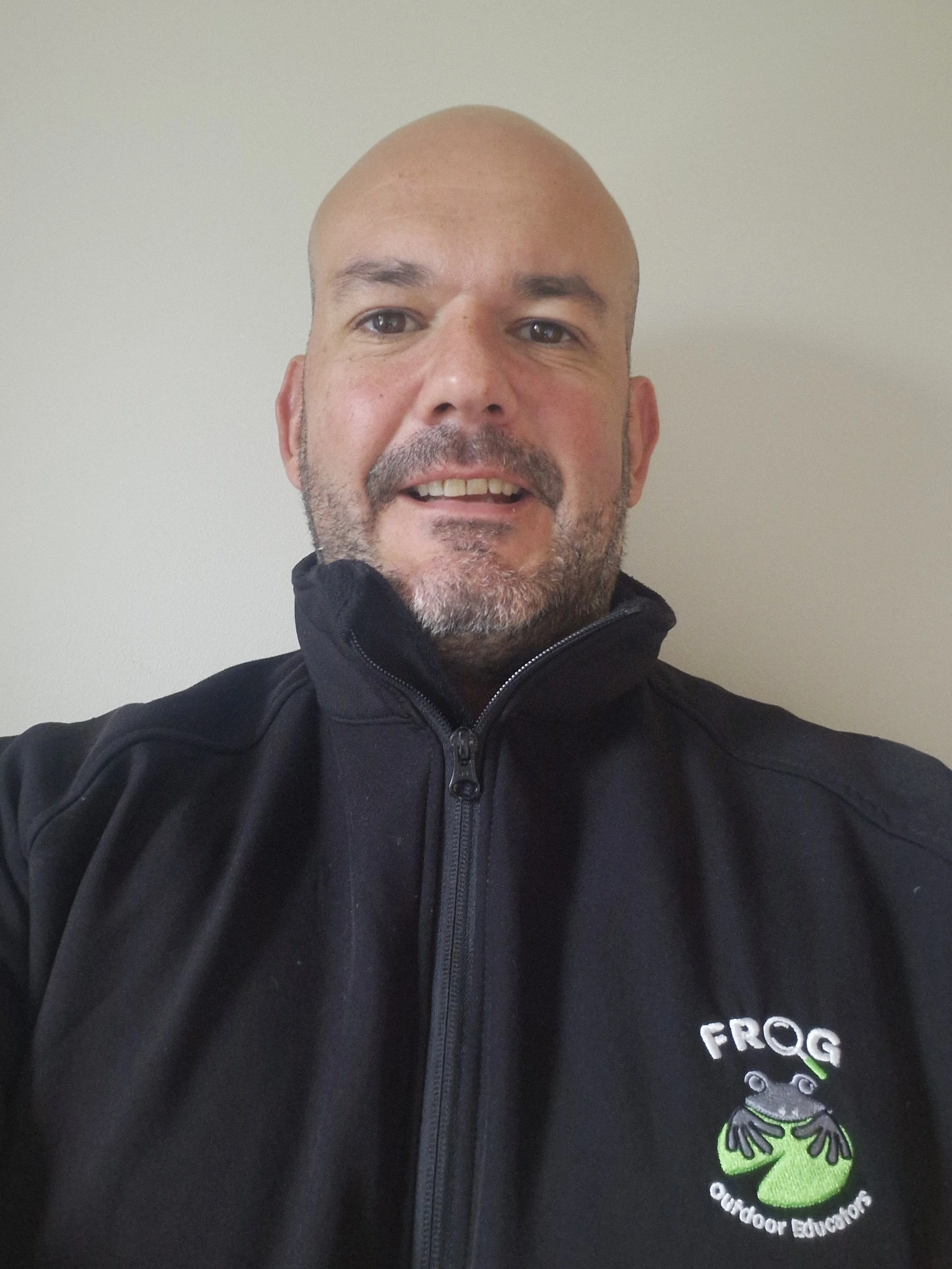 A man taking a selfie wearing a black jacket with a Frog Outdoor Educators logo on the chest.