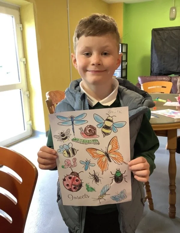 A young boy holding a drawing of various insects and butterflies, smiling inside a room with green and yellow walls.