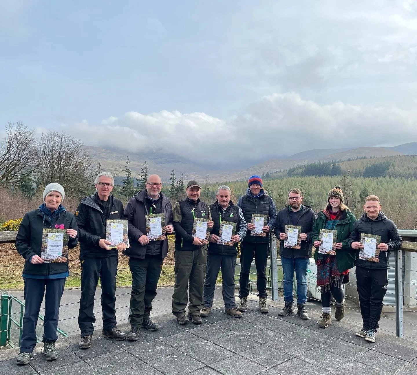 A group of nine people standing outdoors on a balcony, each holding a certificate, with a scenic background of mountains and trees.