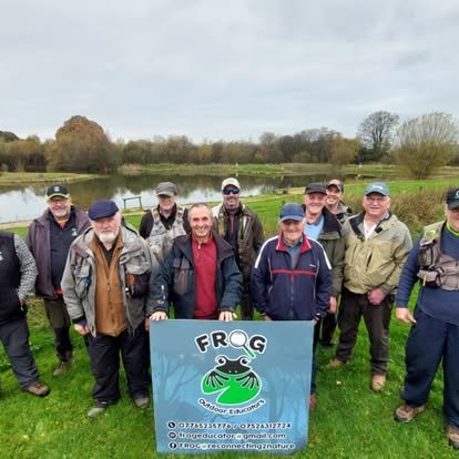 Group of ten men standing outdoors on grass near a lake, holding a sign that reads 'FROG' with frog logo, during daytime, dressed in casual outdoor clothing.
