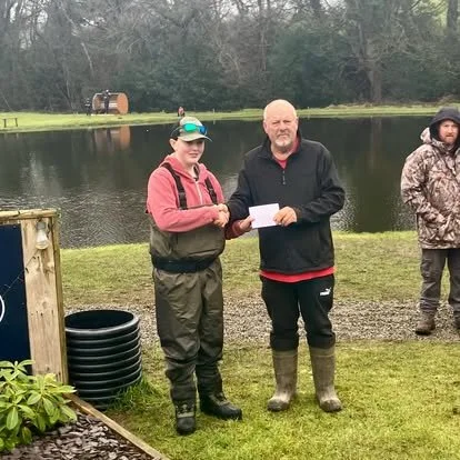Two men standing by a pond; one is giving an award or certificate to the other. Another person stands nearby on the right. Scene takes place outdoors on a grassy area with trees in the background.