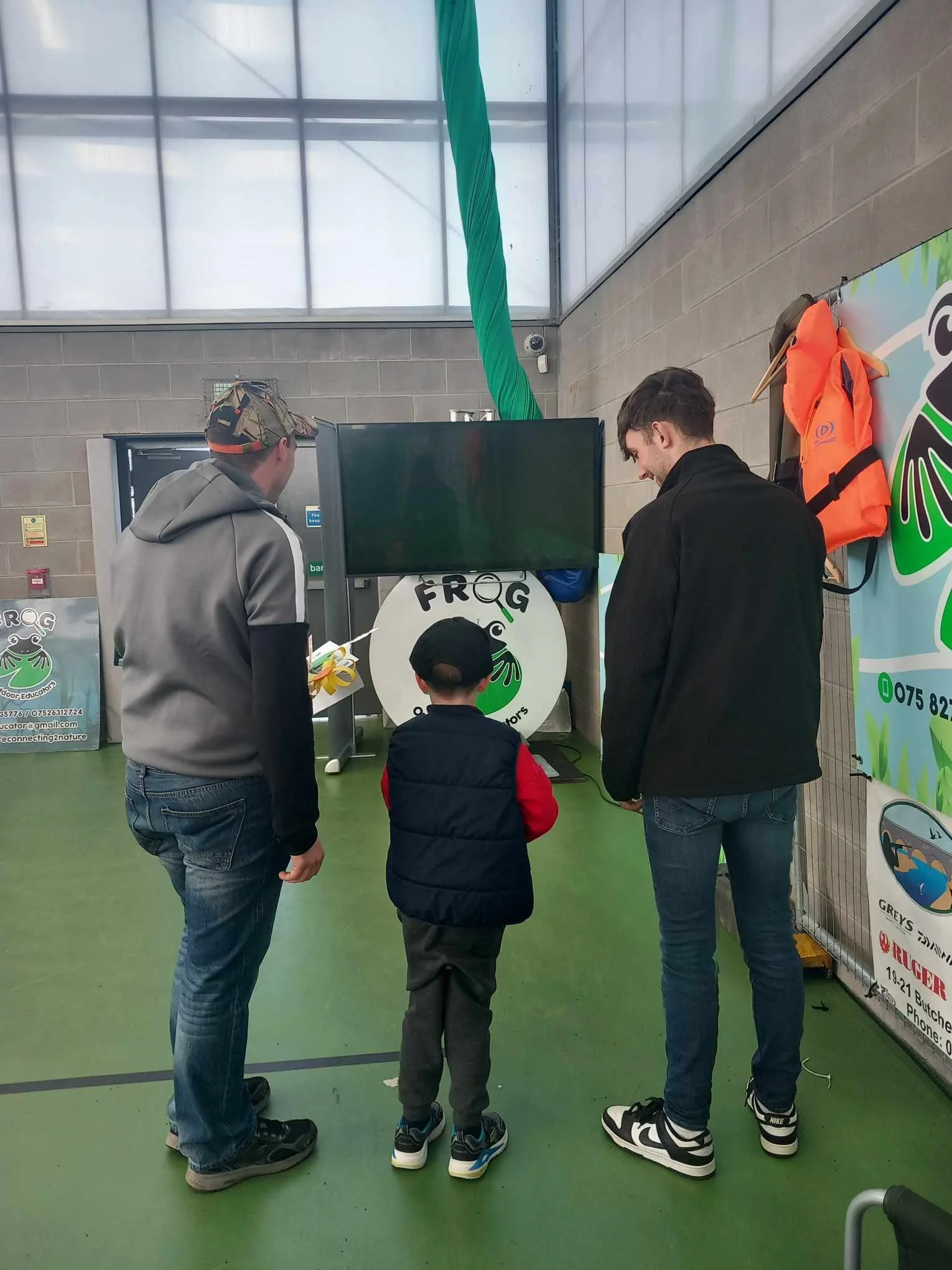 Two men and a young boy stand together indoors, facing a large TV screen. The wall has a large badge with a frog logo and some informational posters. The setting appears to be a recreational or educational facility.