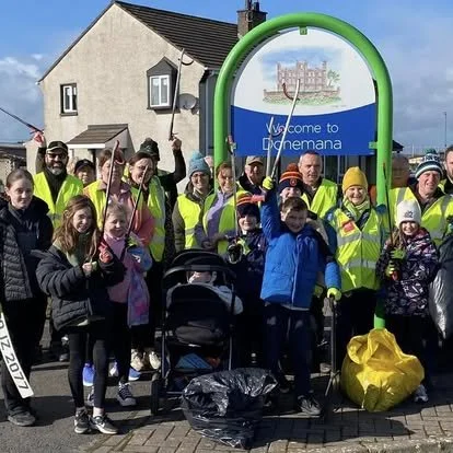 Group of people, including children and adults, wearing yellow safety vests, gathered at a bus stop in Dromena, Ireland, with some holding pink flags and a stroller in front, under a sign that says 'Welcome to Dromena'.