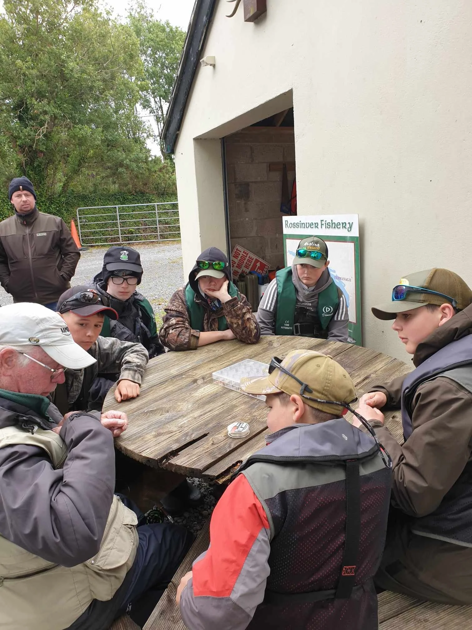 A group of people, mostly young boys and two adults, are gathered around a large wooden picnic table outside near a building. Some are wearing fishing gear, including hats and vests, indicating a fishing activity, at the Rossinver Fishery. An older man appears to be explaining or instructing, while others listen attentively.