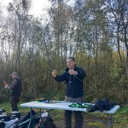 A man standing outdoors behind a table set up with various camera equipment, holding a golf tee, with trees in the background.