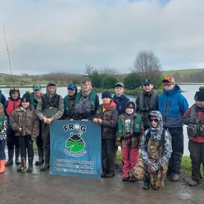Group of people outdoors holding a blue sign with a frog logo and social media information, in a rural area near water and hills.