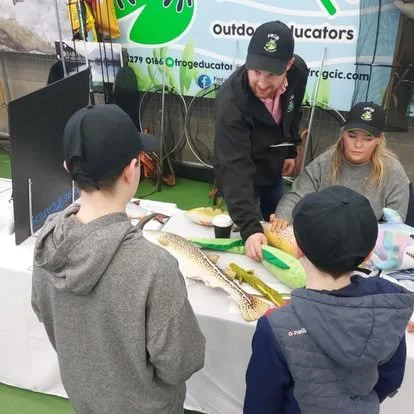A man showing fishing equipment and a fish to three children at an outdoor education booth.