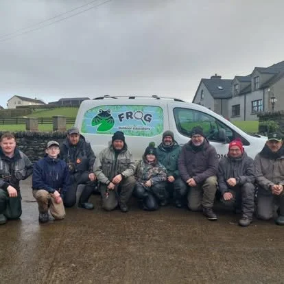 Group of nine people kneeling in front of a white vehicle with a frog mascot logo and the words "FRQ" and "Outdoor Education" on it, on a muddy area near residential houses.
