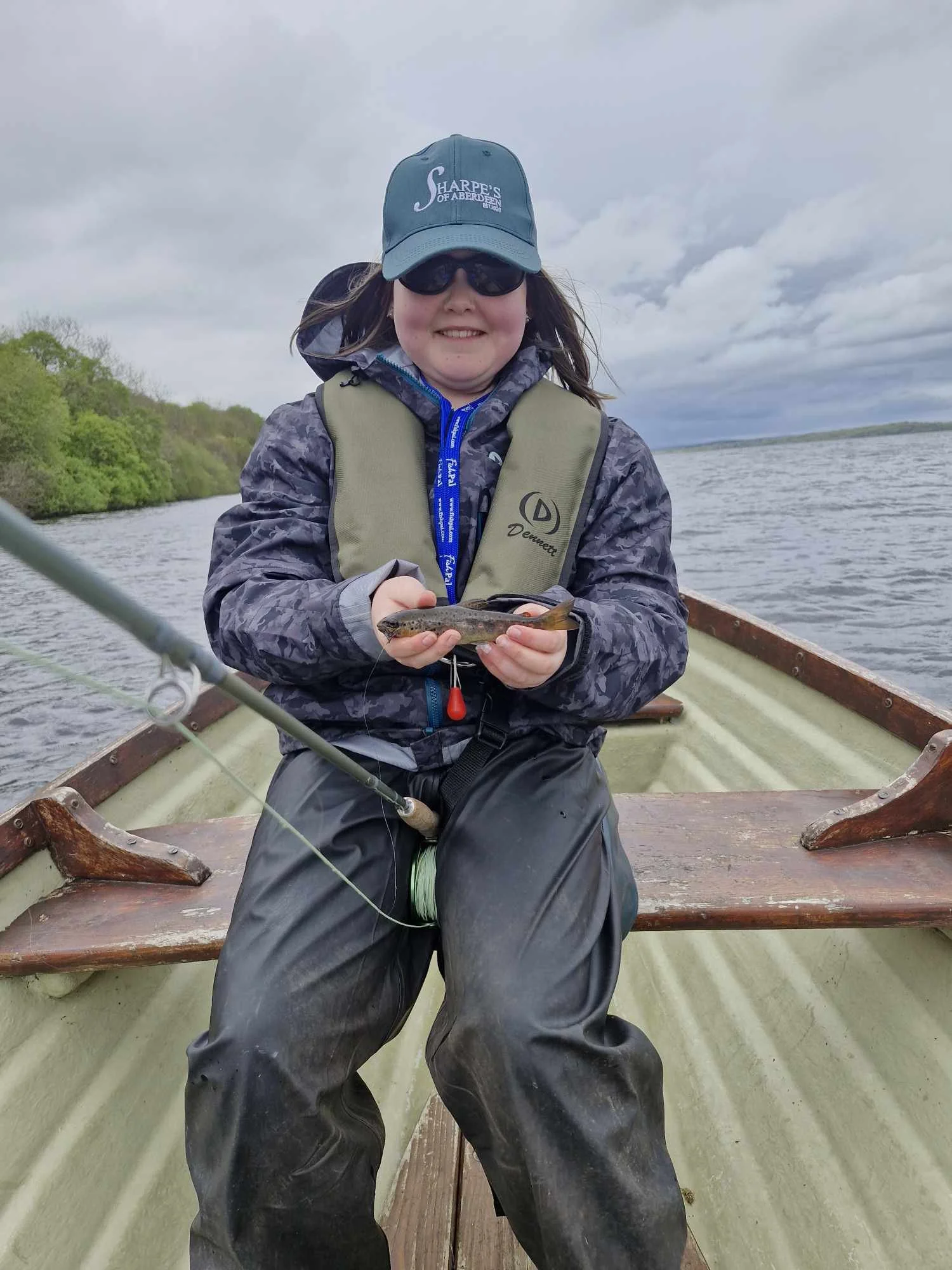 Young girl in a boat holding a small fish, wearing a cap, sunglasses, and rain gear, with a lake and cloudy sky in the background.