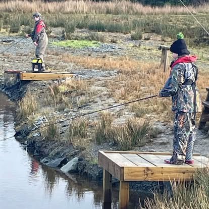Two children wearing camouflage clothing and winter hats fishing on wooden docks near a marshy area.