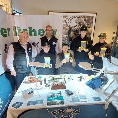 Group of five adults and children holding yellow boxes at a table with various electronic equipment and pamphlets, indoor setting with a banner in the background.